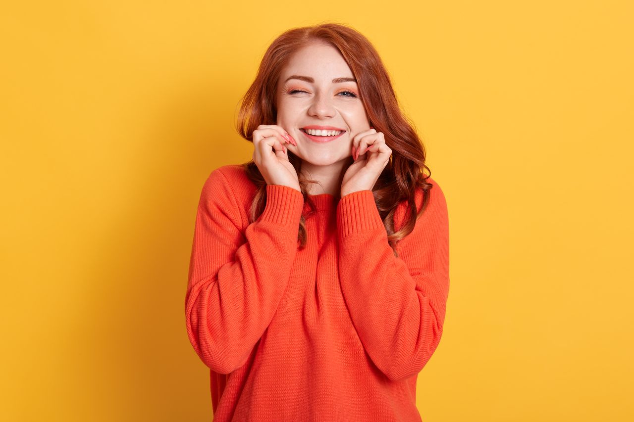 Jeune femme pétillante aux cheveux blonds attachés, pull coloré, sourire malicieux, portrait sur fond jaune doux