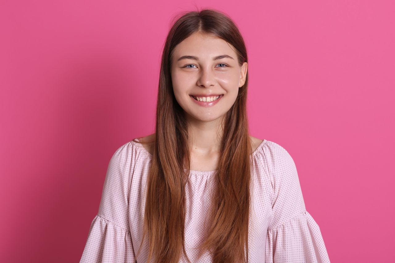 Jeune femme souriante aux cheveux bruns ondulés, teint lumineux, portrait beauté sur fond rose poudré